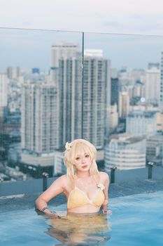 Cosplay model in a yellow bikini posing in a rooftop pool with a stunning cityscape backdrop.