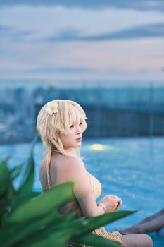 Blonde woman relaxing by a rooftop pool with cityscape during sunset, flower in hair.