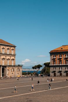 A bustling piazza in Naples with historical architecture and a stunning seaview in the background.