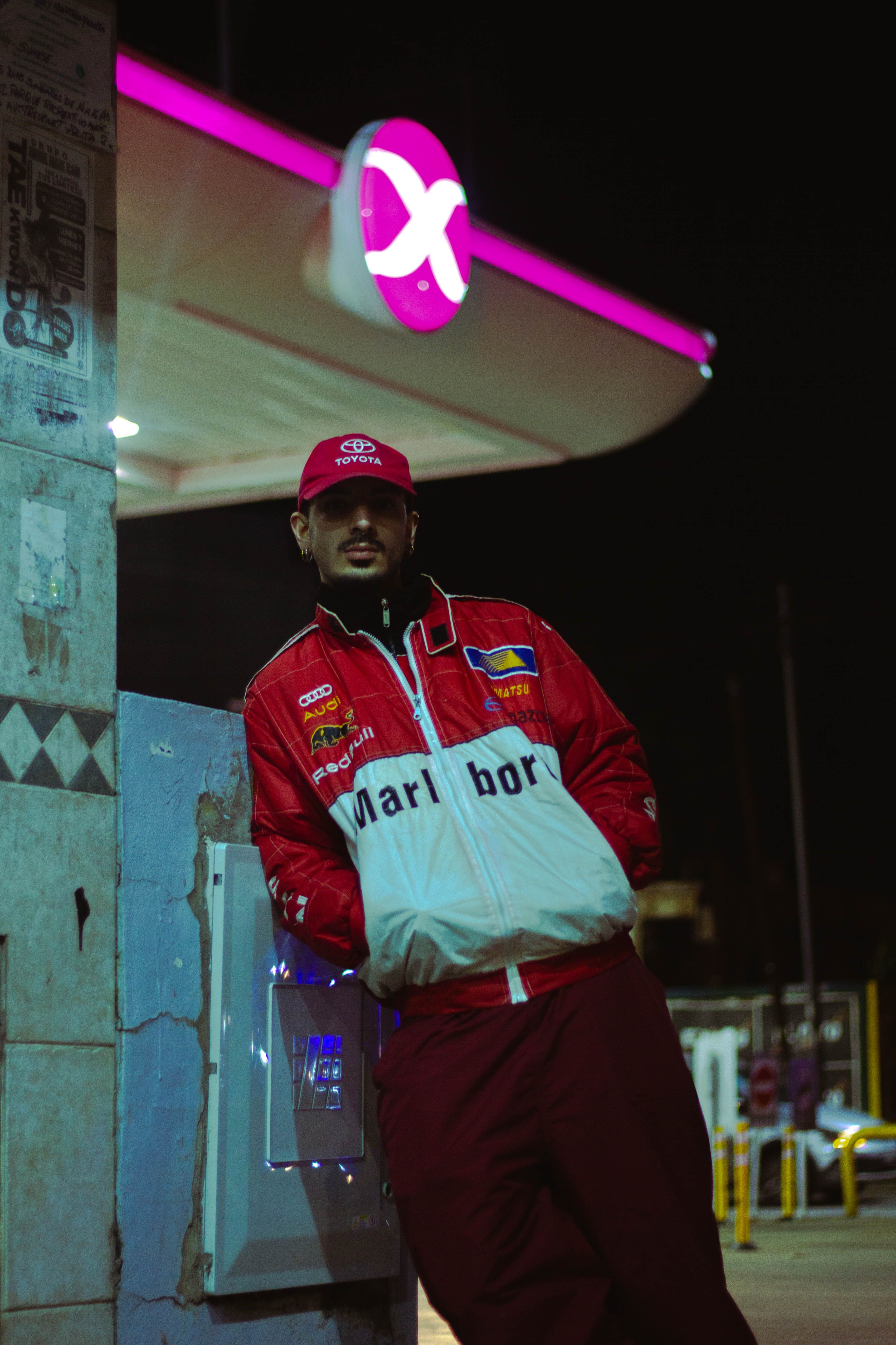 Man in racing jacket leans against wall at an Argentinian gas station at night.