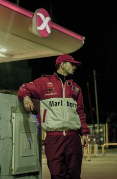 A vintage racecar driver standing at a gas station in Argentina at night, exuding retro vibes.