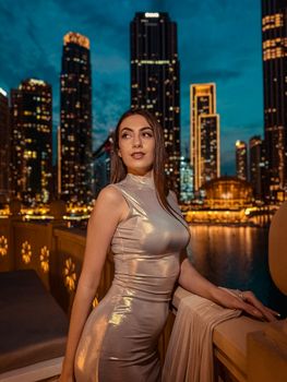 Woman in shimmering dress poses on Dubai balcony with city skyline at night.