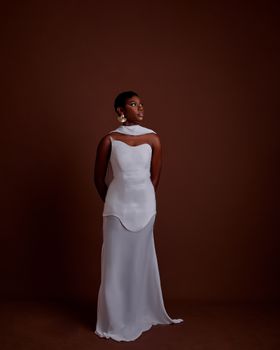 A poised woman in a white dress stands elegantly against a brown background, exuding grace.