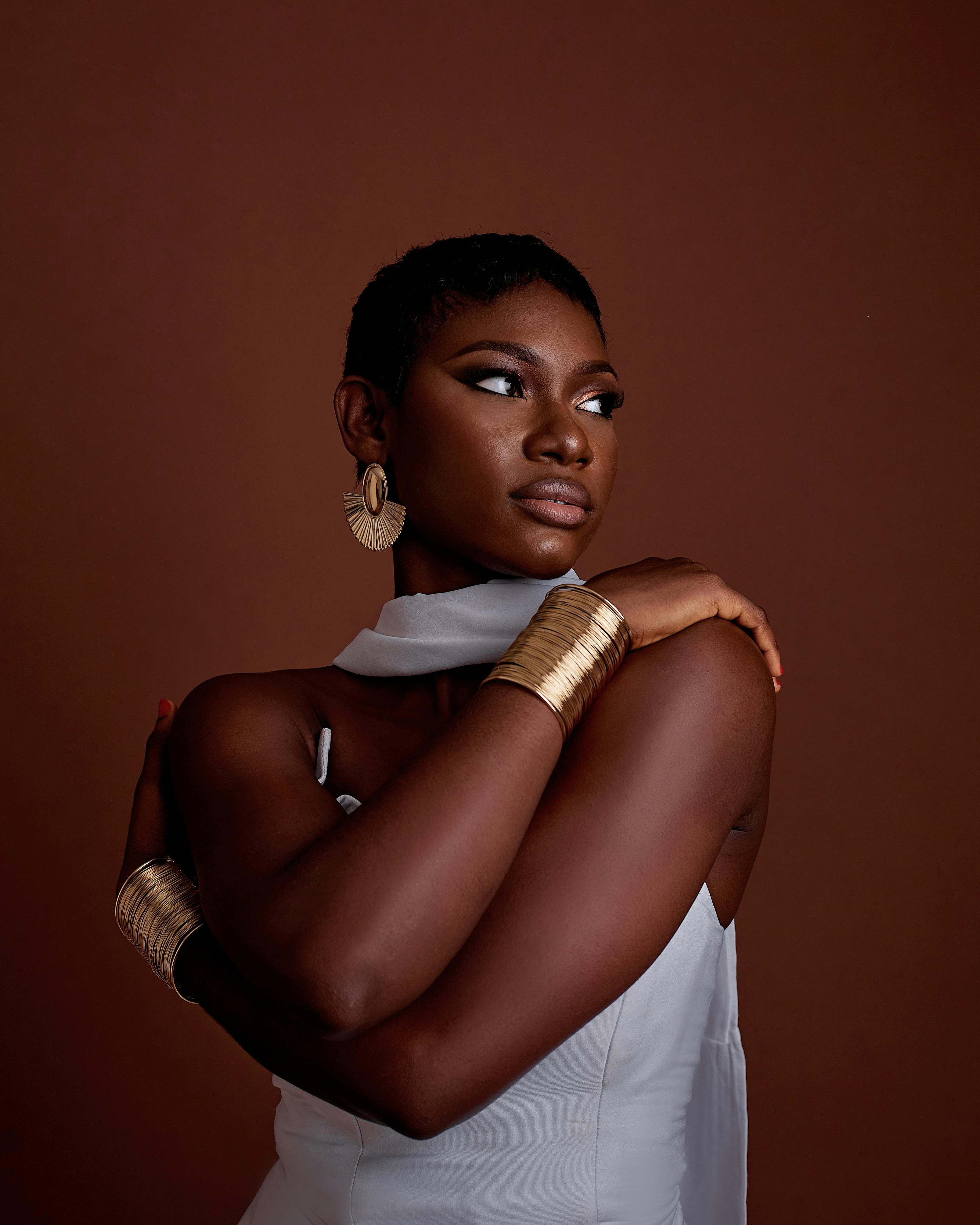 Gratis Stilig kvinne i hvit kjole med gullsmykker mot brun bakgrunn. Arkivbilde
