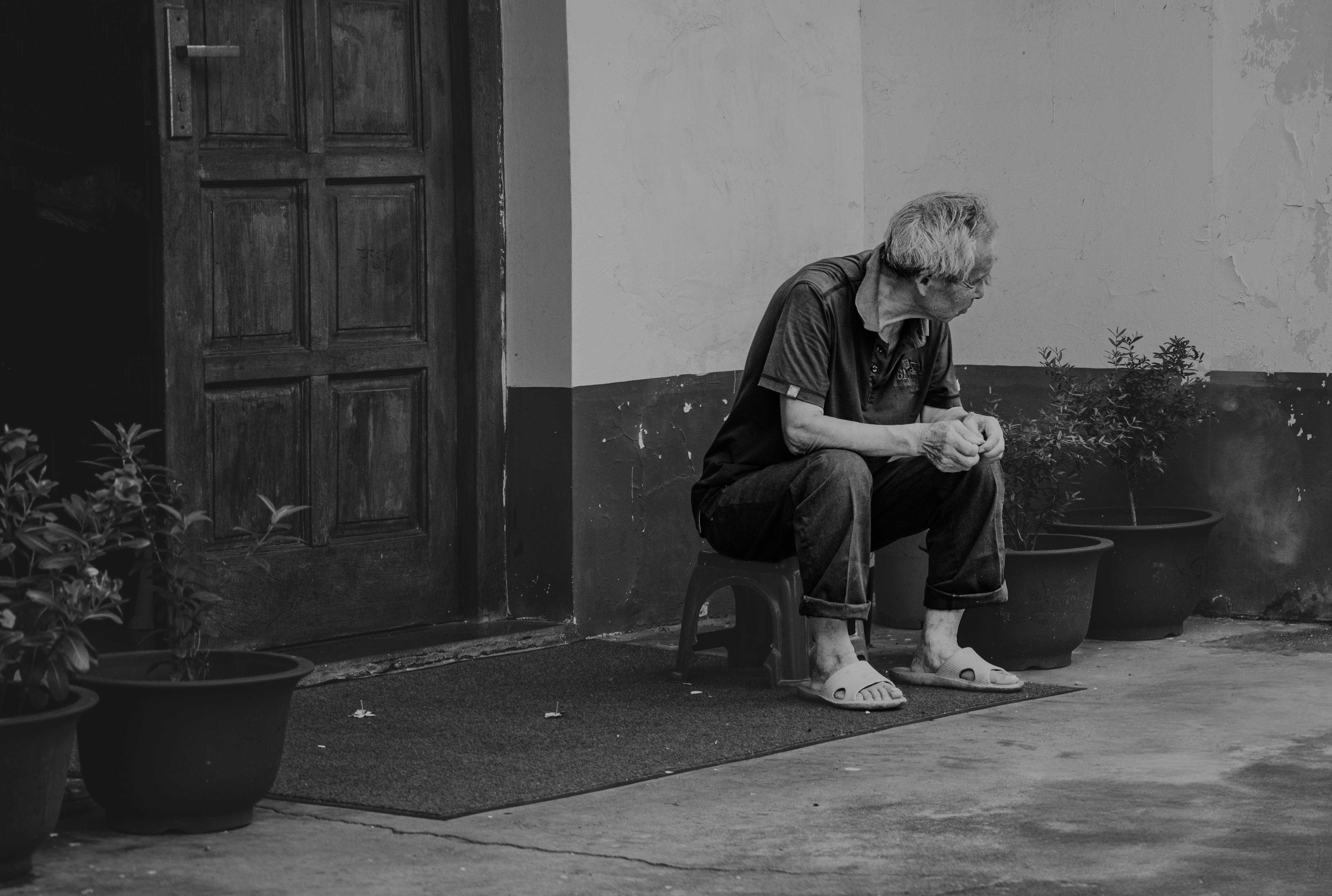 A senior man sits alone on a stool outside a door in a black and white photograph.