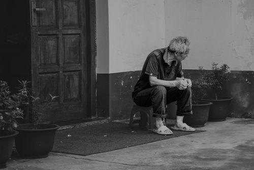Free A senior man sits alone on a stool outside a door in a black and white photograph. Stock Photo