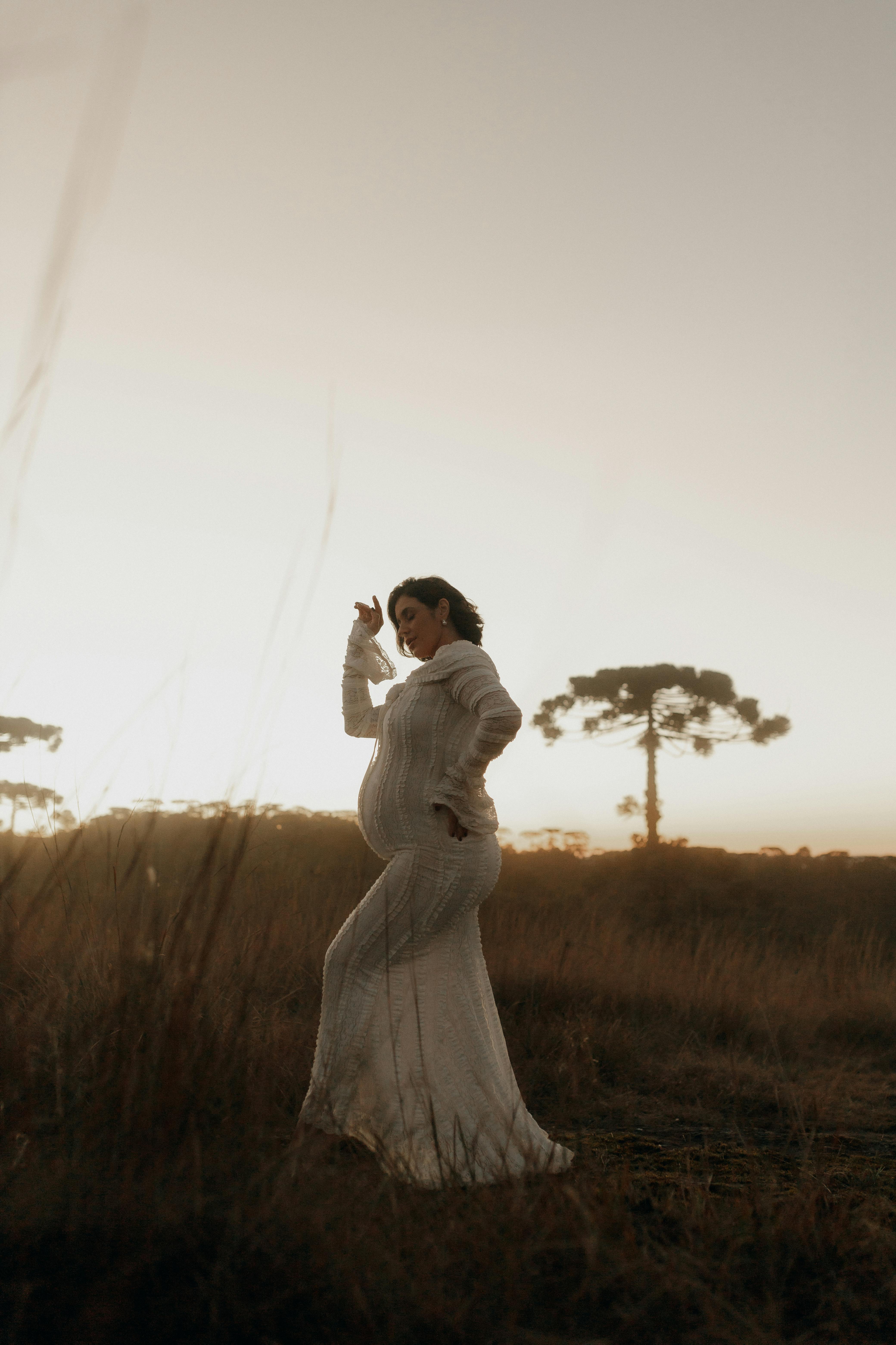 免費 日落時分，身穿白色連身裙的孕婦在寧靜的田野中優雅地擺姿勢。 圖庫相片