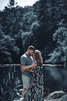 A loving couple shares an intimate moment by a peaceful lake surrounded by lush forest.