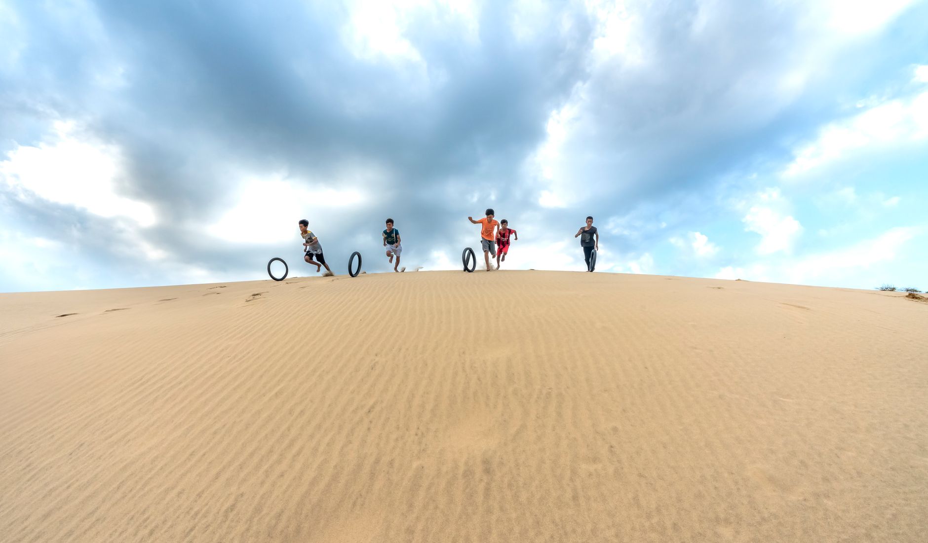 Five children running down sand dunes while playing with tires under a cloudy sky.