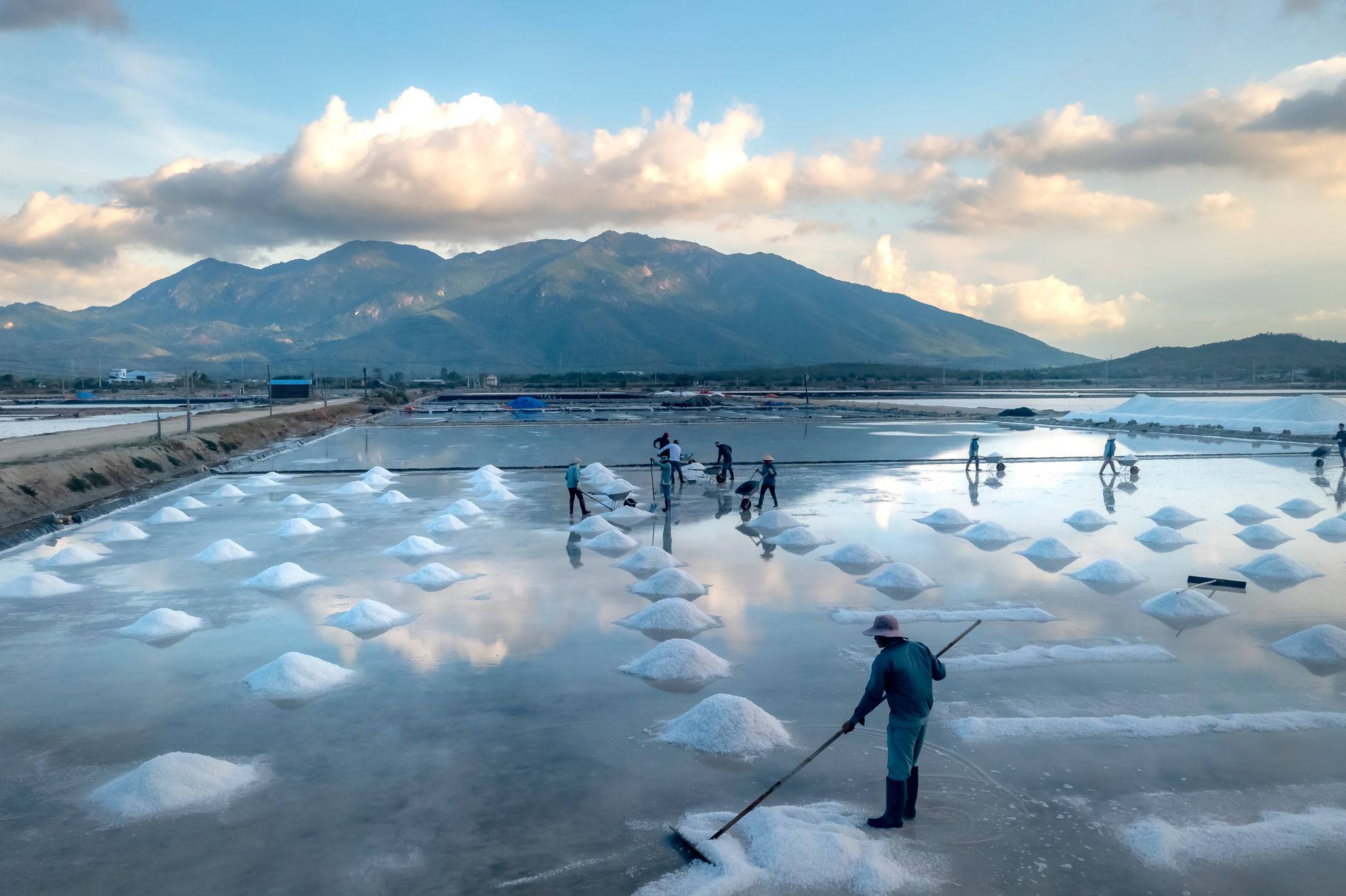 Workers harvesting salt in coastal flats against picturesque mountain backdrop.