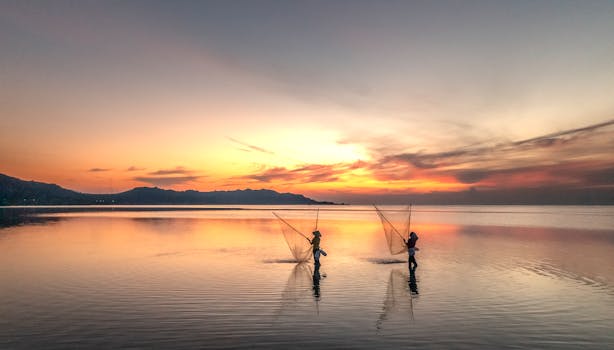 Two fishermen casting nets at sunrise on a calm lake with colorful sky.