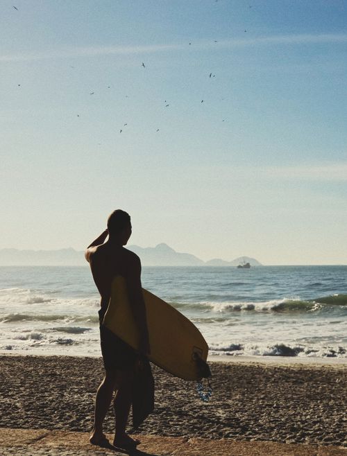 Free A lone surfer holding a surfboard looks out over the ocean waves at a sandy beach under a clear sky. Stock Photo