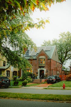 A picturesque brick house with solar panels, lush greenery, and fall foliage in a suburban neighborhood.