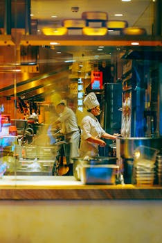 Chefs preparing meals in a vibrant Asian restaurant kitchen, seen through the glass.