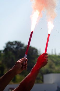 Two hands holding red flares with smoke on a sunny day outdoors, trees in the background.