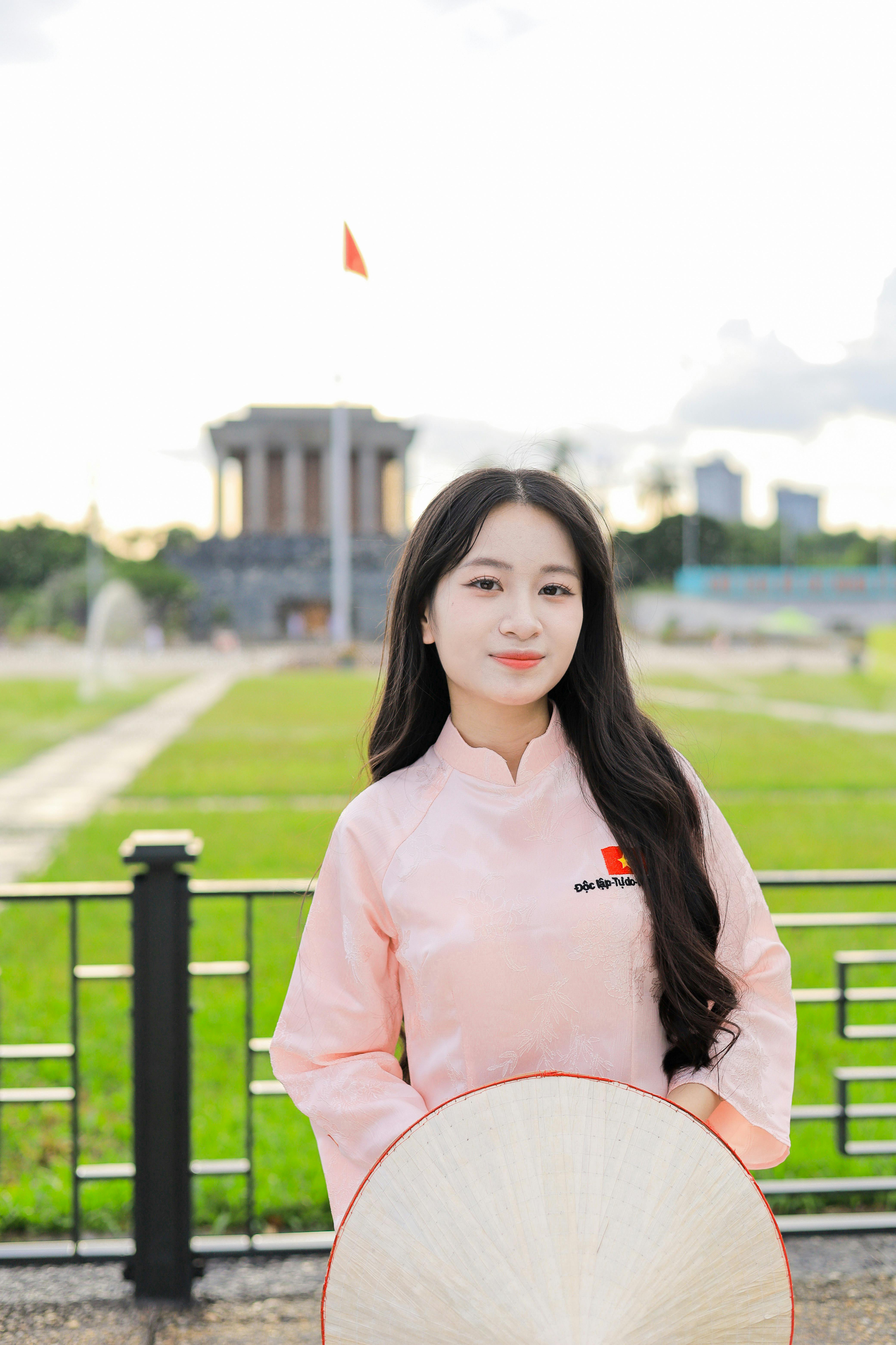 A woman in pink áo dài smiles, standing near the Ho Chi Minh Mausoleum in Vietnam.