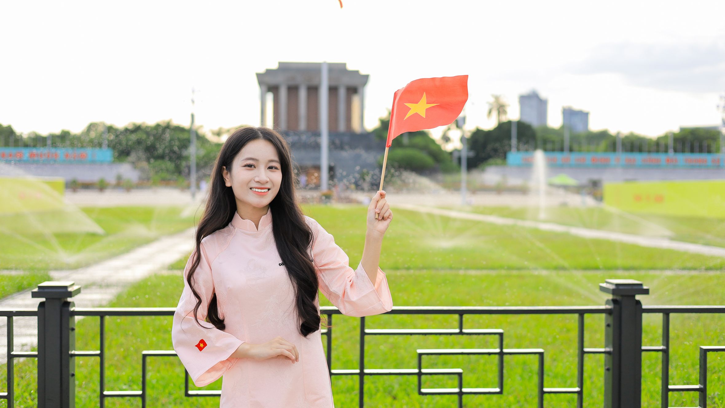 Young woman in traditional dress holding Vietnamese flag in front of a famous landmark in Hanoi.