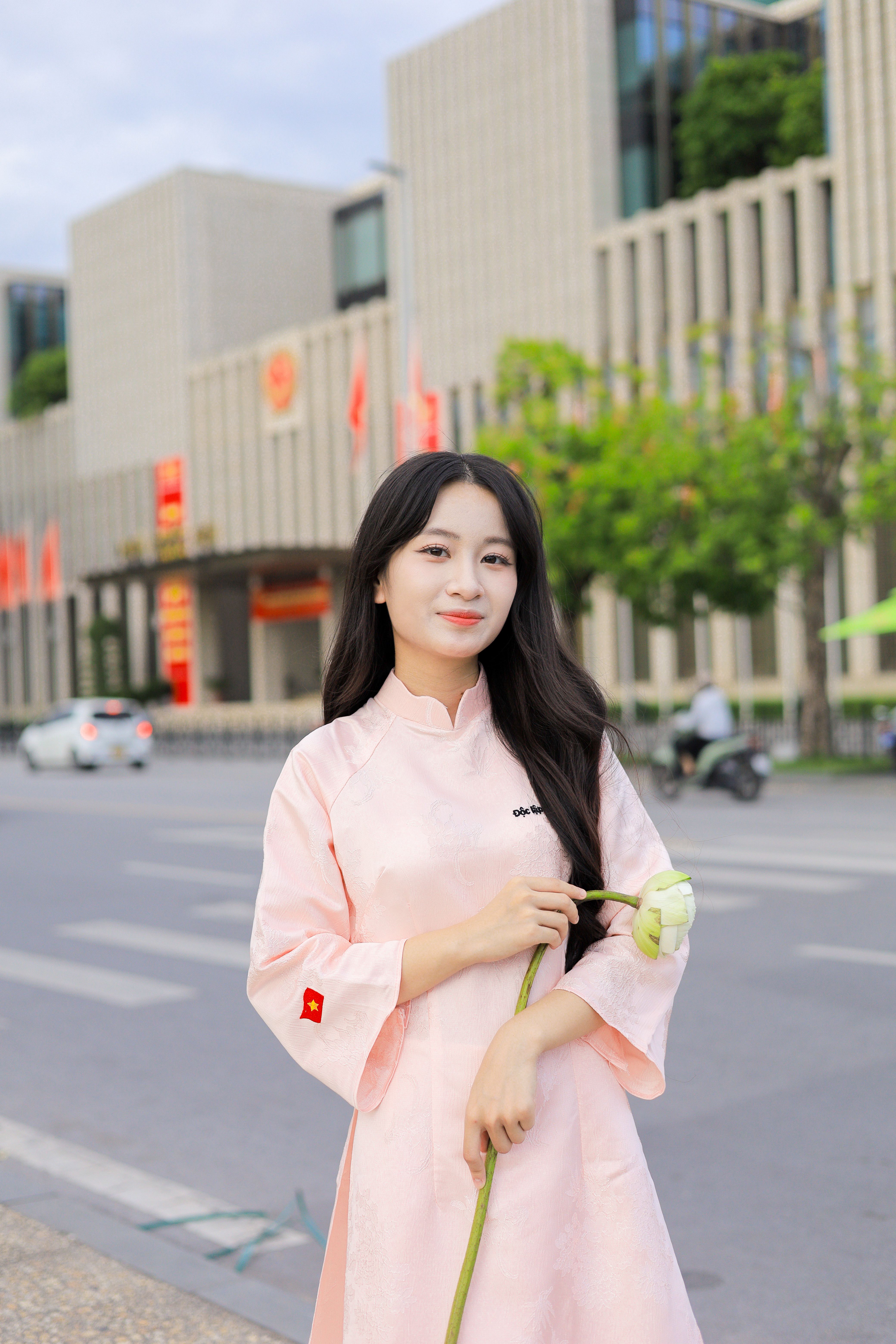 A young woman in traditional Vietnamese attire holds a lotus flower in an urban setting.