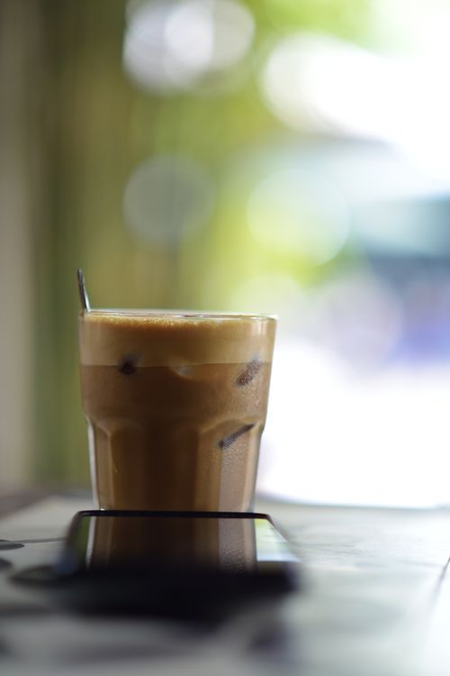 Free Refreshing iced coffee in a glass on a table, with a blurred smartphone in a sunny cafe. Stock Photo