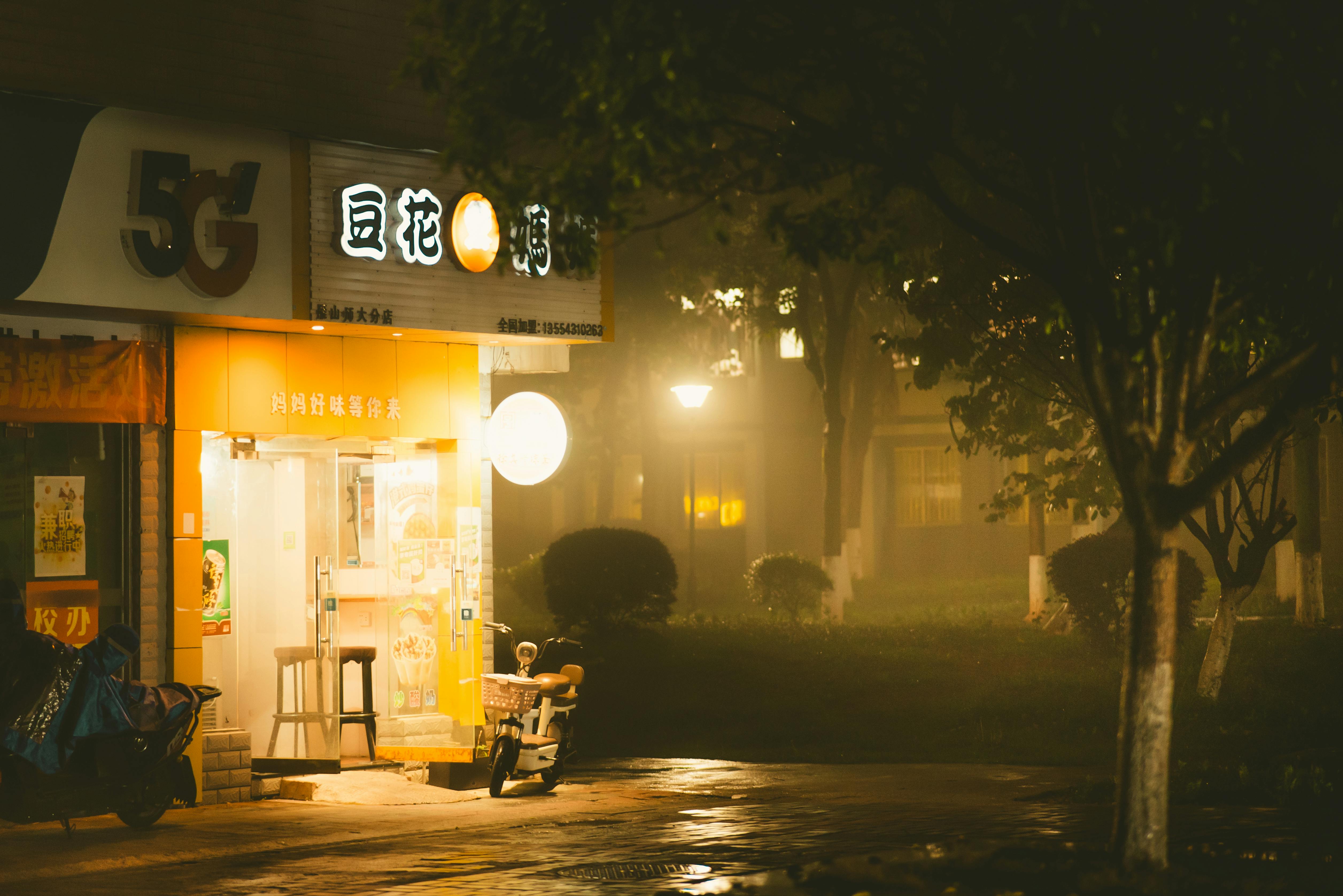 A misty evening view of a warmly lit Chinese street shop with scooters parked outside.