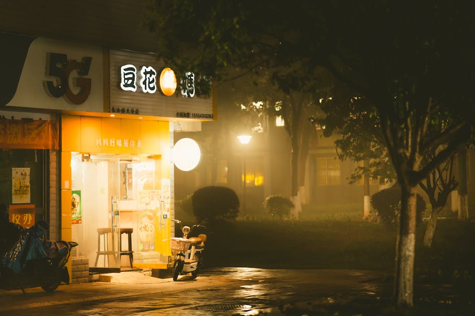 A misty evening view of a warmly lit Chinese street shop with scooters parked outside.