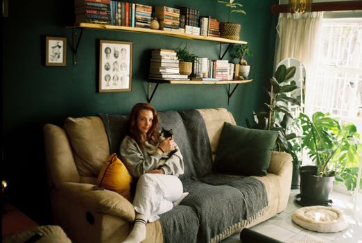Woman relaxing with a cat in a cozy living room full of books and plants.