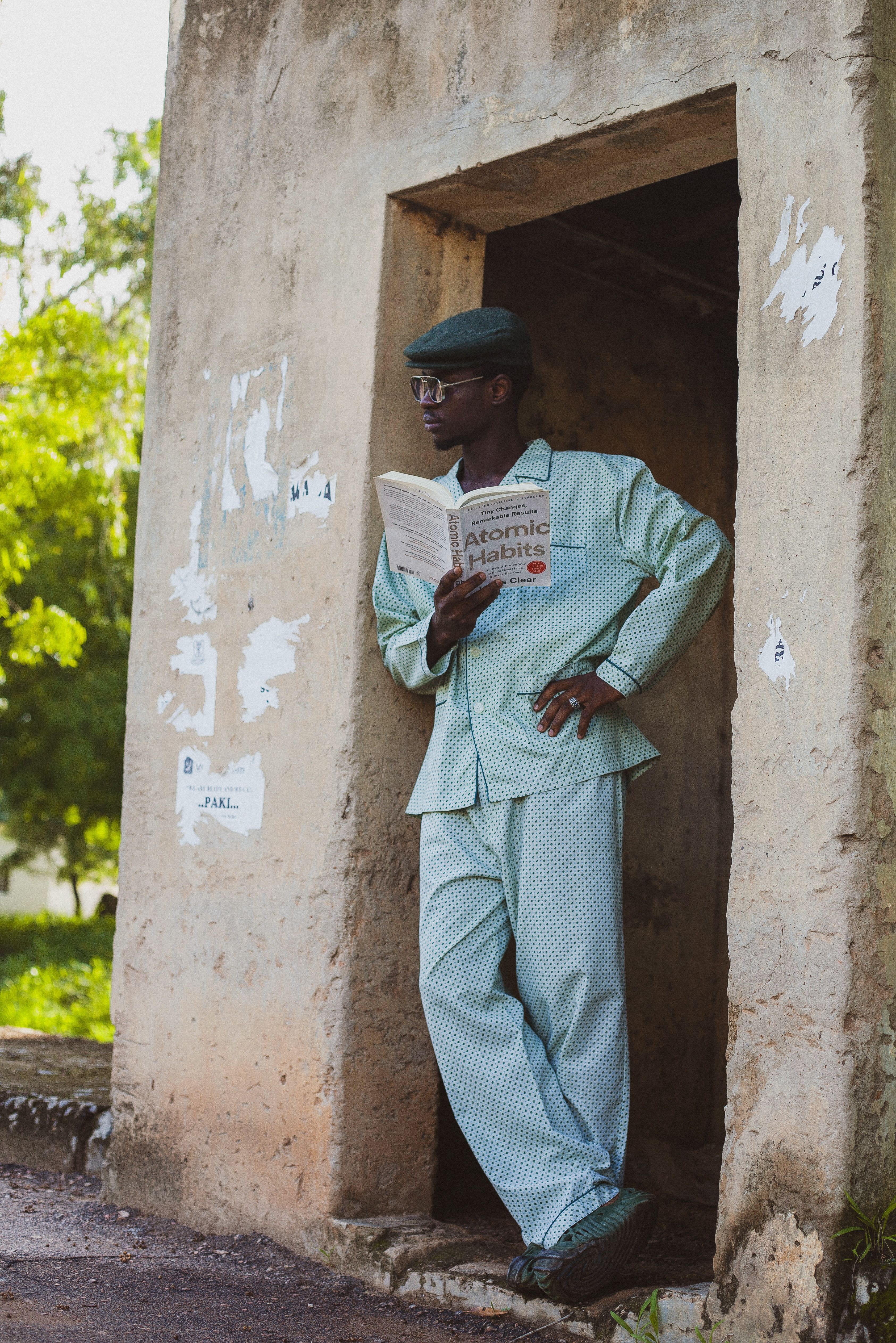 A man in stylish clothing leans in a doorway reading 'Atomic Habits' outdoors.