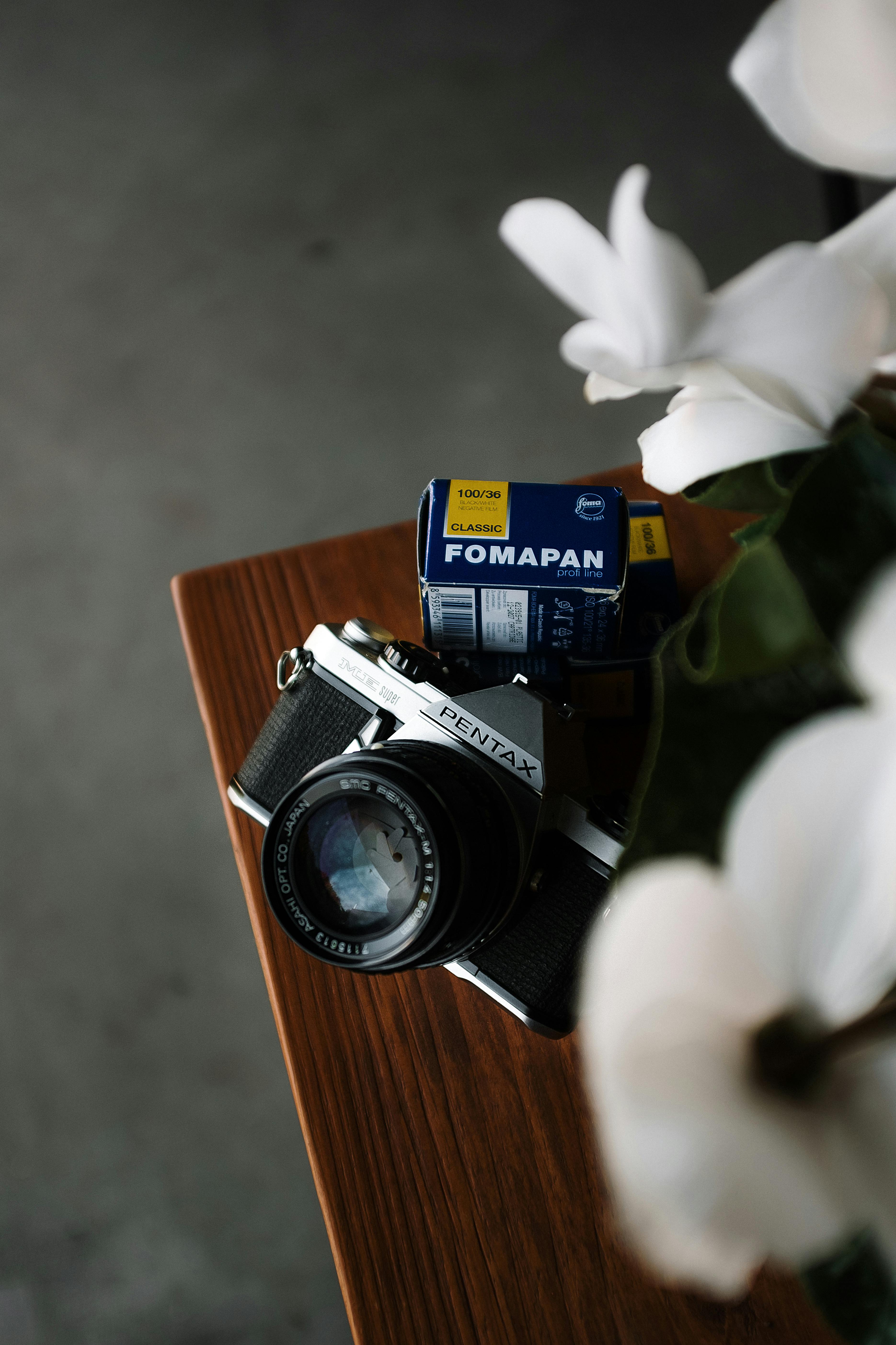 A classic Pentax camera with Fomapan film beside white flowers, evoking analog nostalgia.
