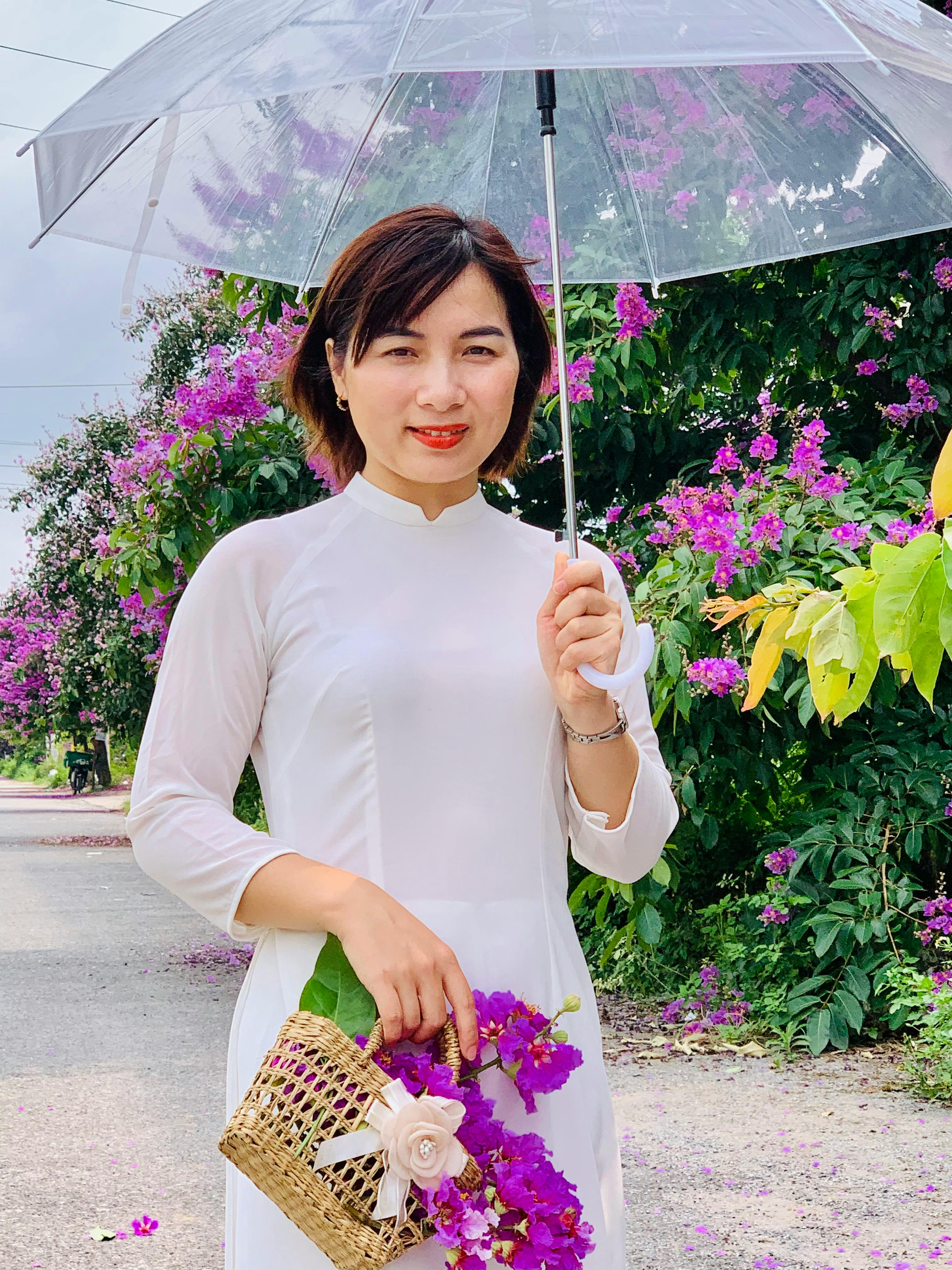 Woman in traditional Ao Dai holding clear umbrella and flowers, smiling outdoors.