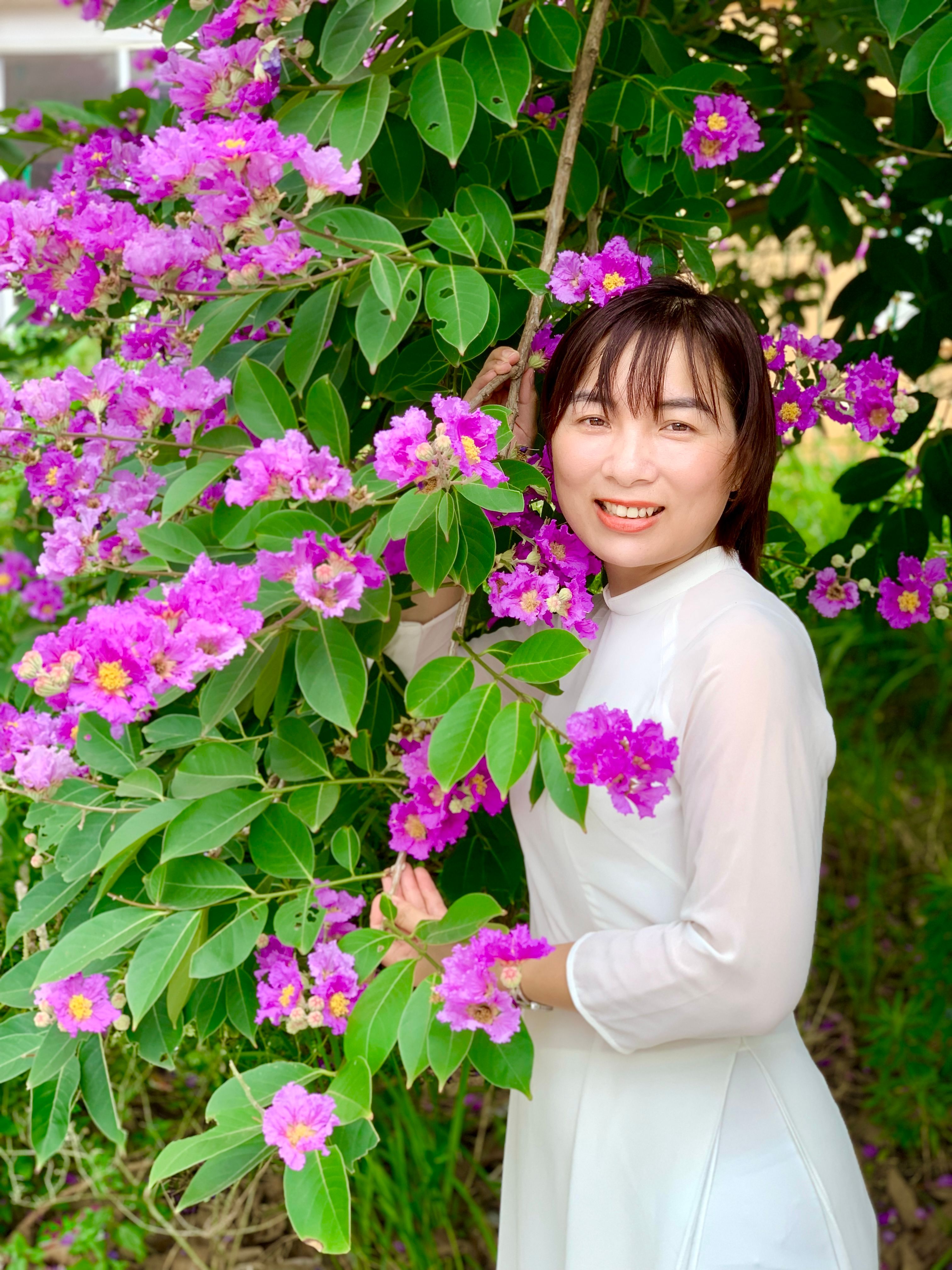 A woman in a white dress smiles beside vibrant purple flowers in Nam Định, Vietnam.