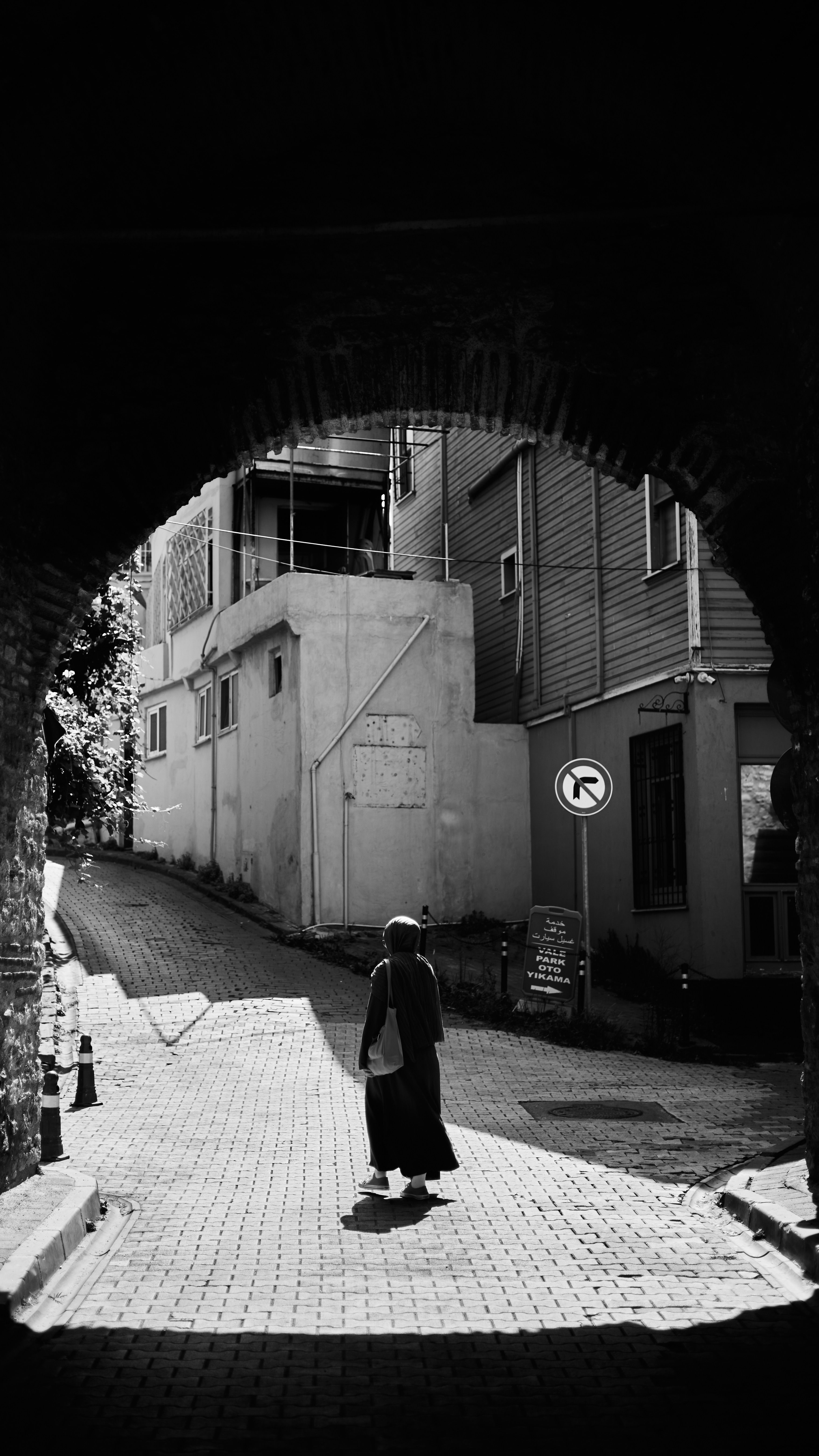 Black and white image of a person walking in a shadowed urban alleyway.