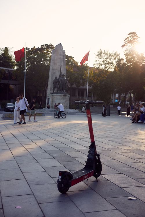 Free A red electric scooter stands in front of a monument in a vibrant city square, capturing sunset hues. Stock Photo