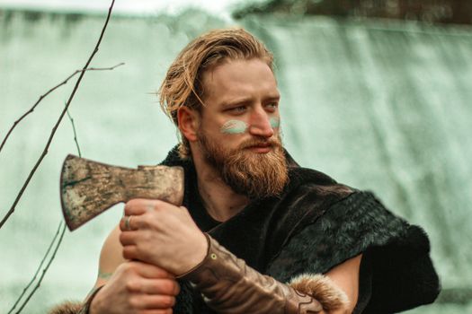 Bearded man dressed as a Viking with face paint, holding an axe outdoors.