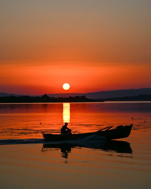 Free A person in a small boat is silhouetted against a stunning sunset over a calm lake, creating a peaceful scene. Stock Photo