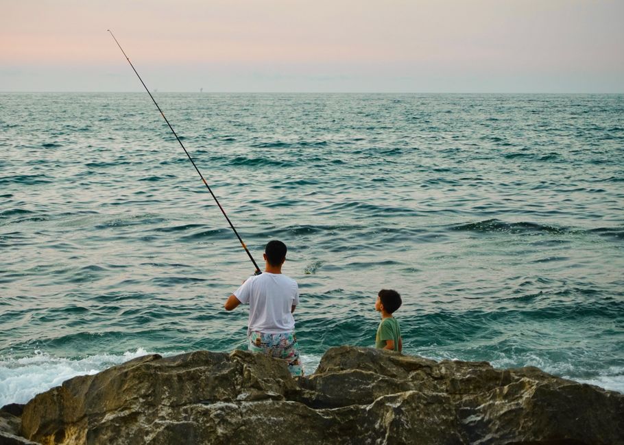 A father and son enjoy fishing on ocean rocks during a serene sunset.