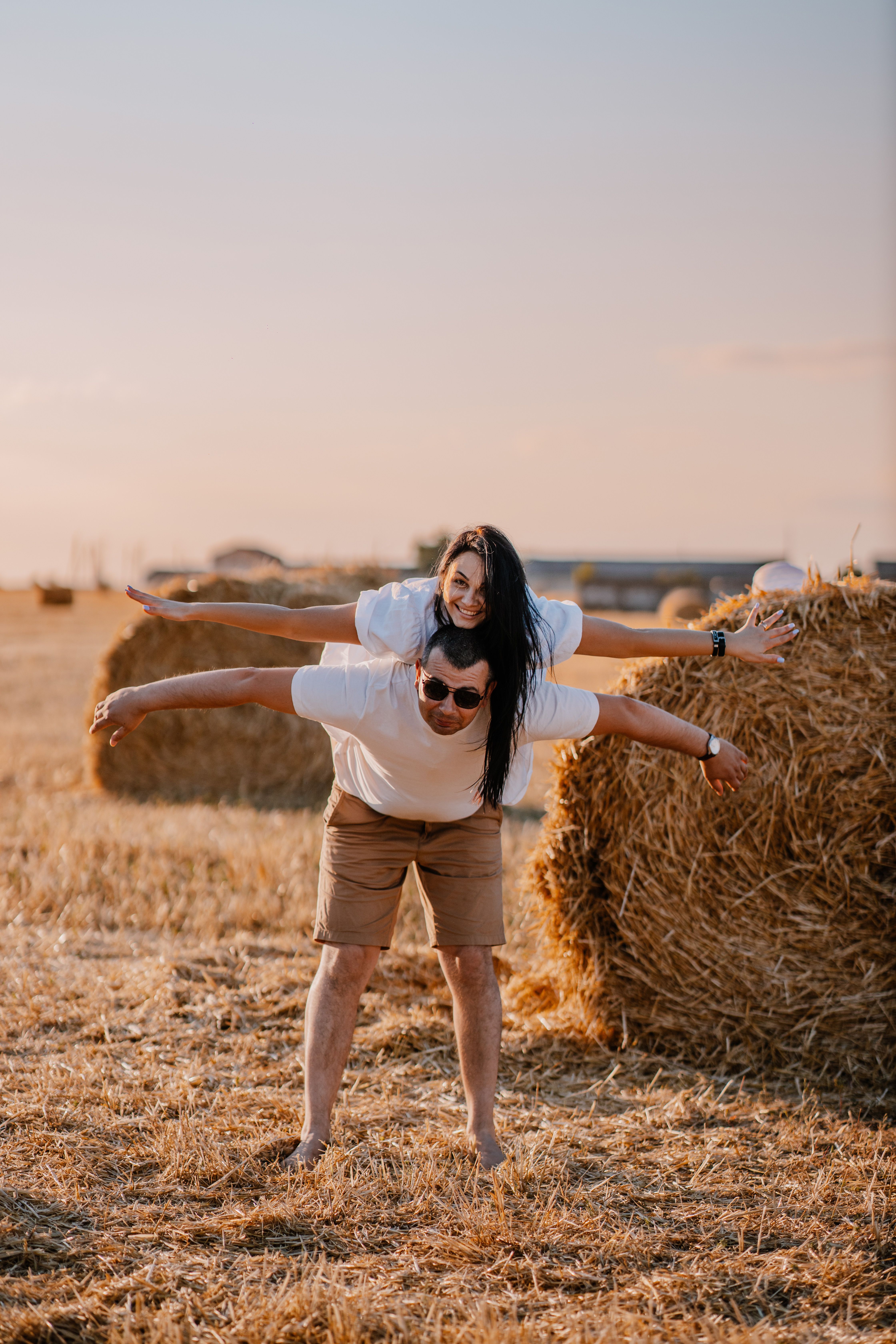Joyful couple enjoying playful moments in a sunny wheat field with hay bales at sunset.