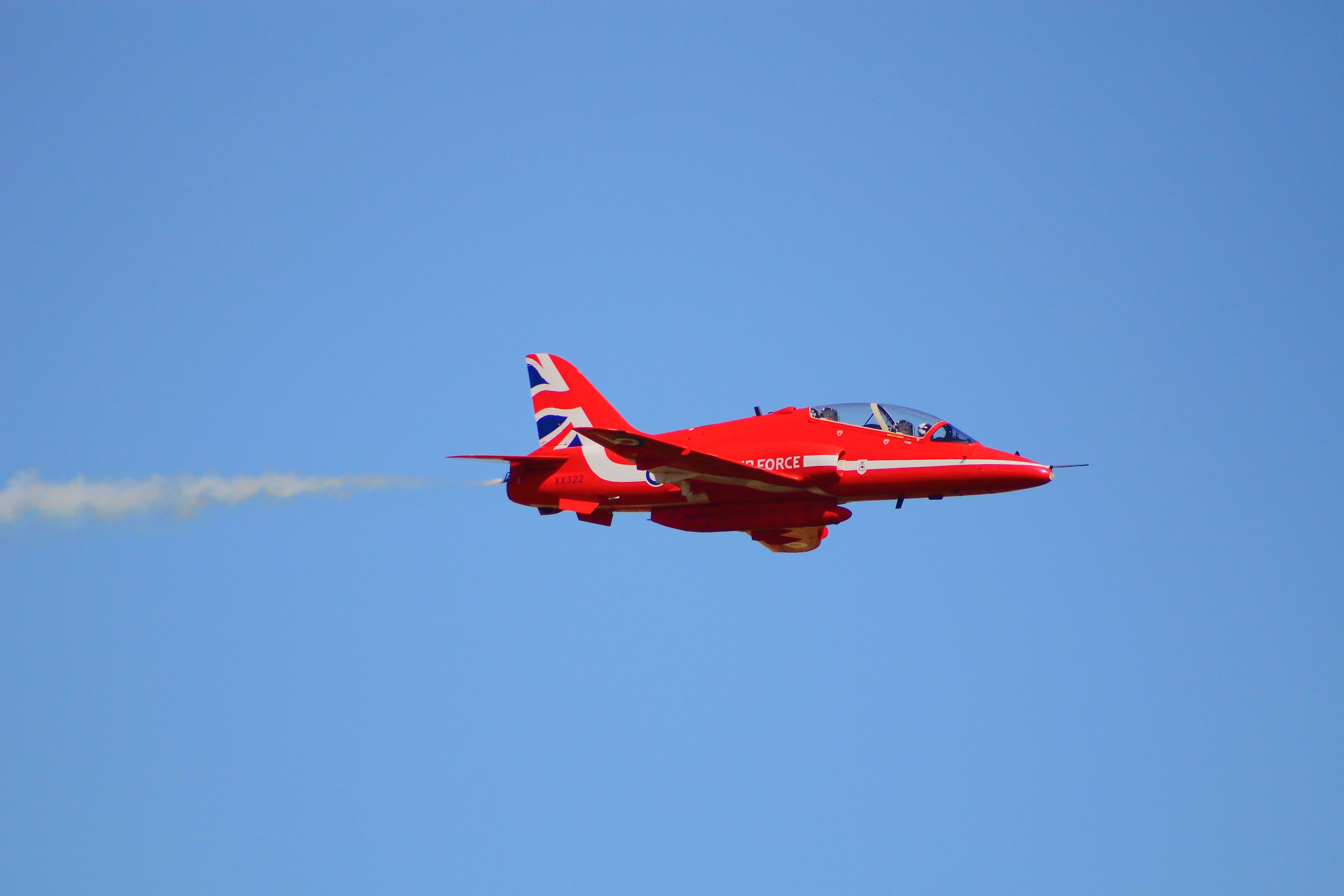 Red jet plane with British flag design soaring through clear blue sky emitting vapor trails.