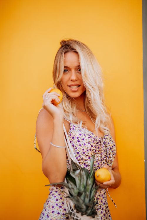 Free Blonde woman in floral dress holding lemons and pineapple against a bright yellow wall, conveying a summer vibe. Stock Photo