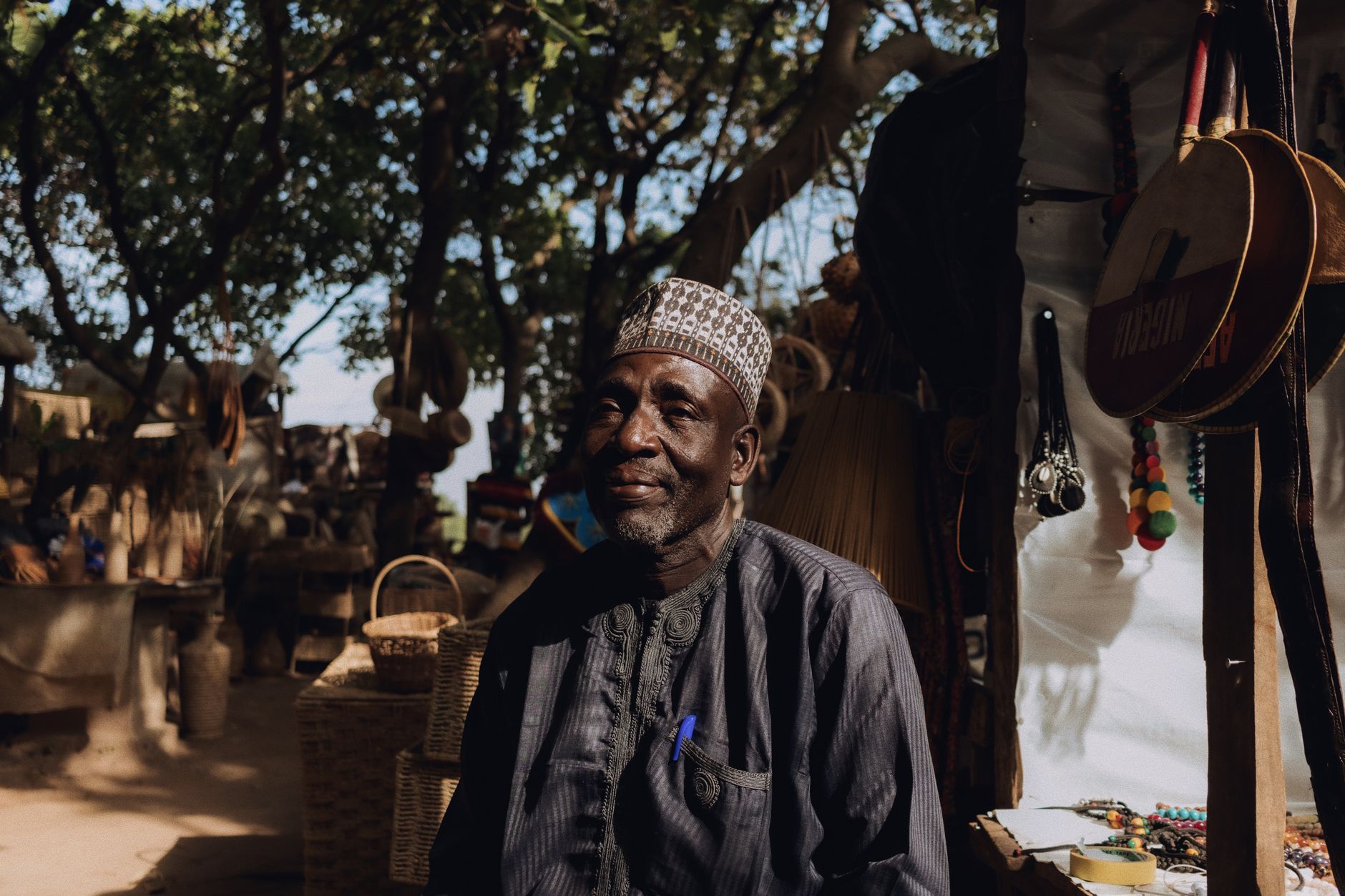 Elderly man at Nigerian market surrounded by traditional crafts and art under warm daylight.