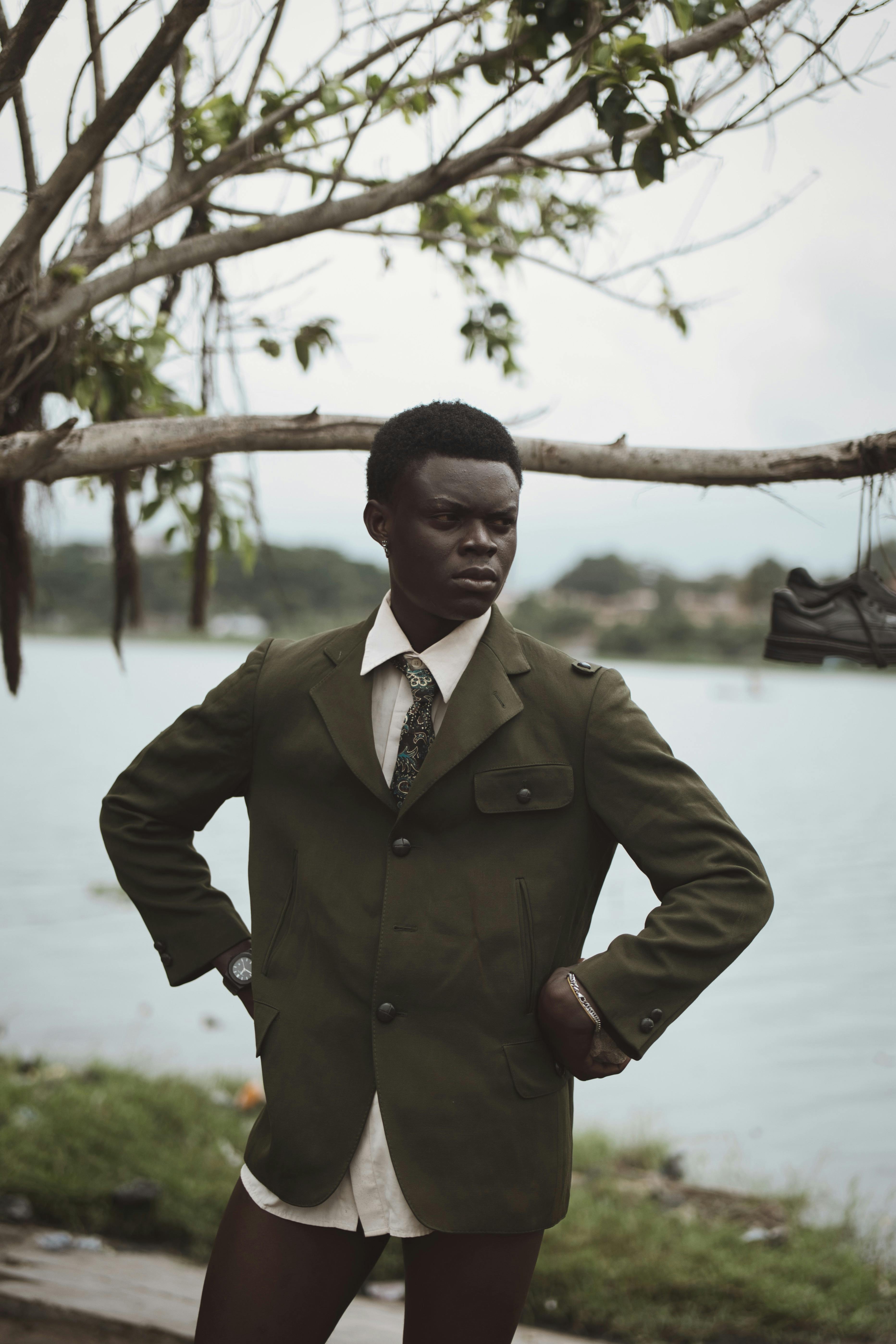 A man in a formal outfit confidently poses by the lake under a tree, conveying elegance and style.