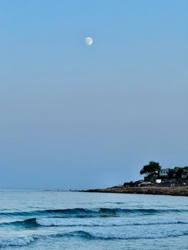Serene view of the moon over Alicante's rocky coastline during twilight with gentle waves.