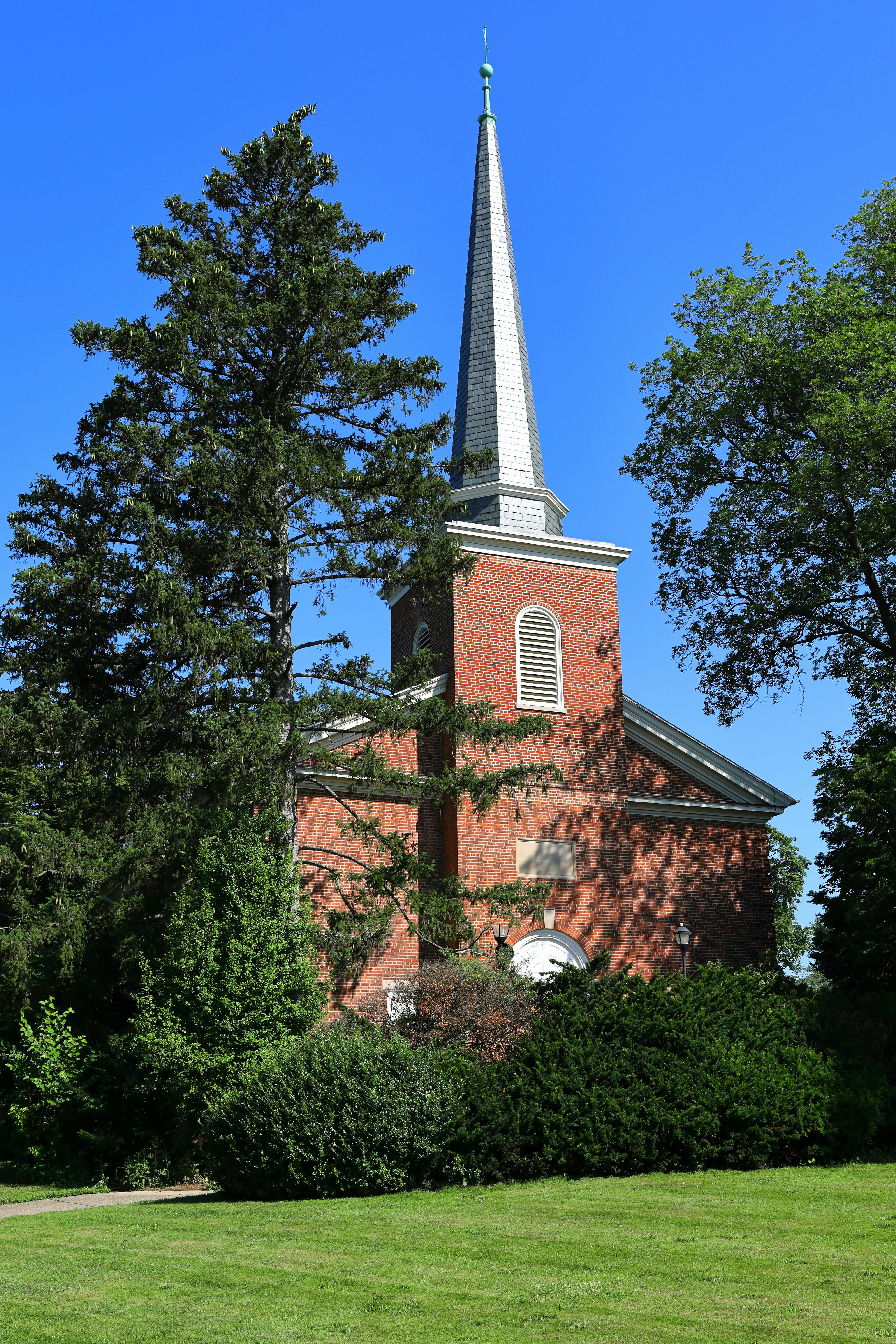 Red brick church with tall steeple surrounded by lush greenery against clear blue sky.