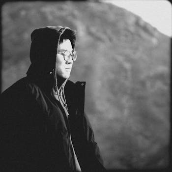 A contemplative person in a hooded jacket outdoors, captured in black and white.