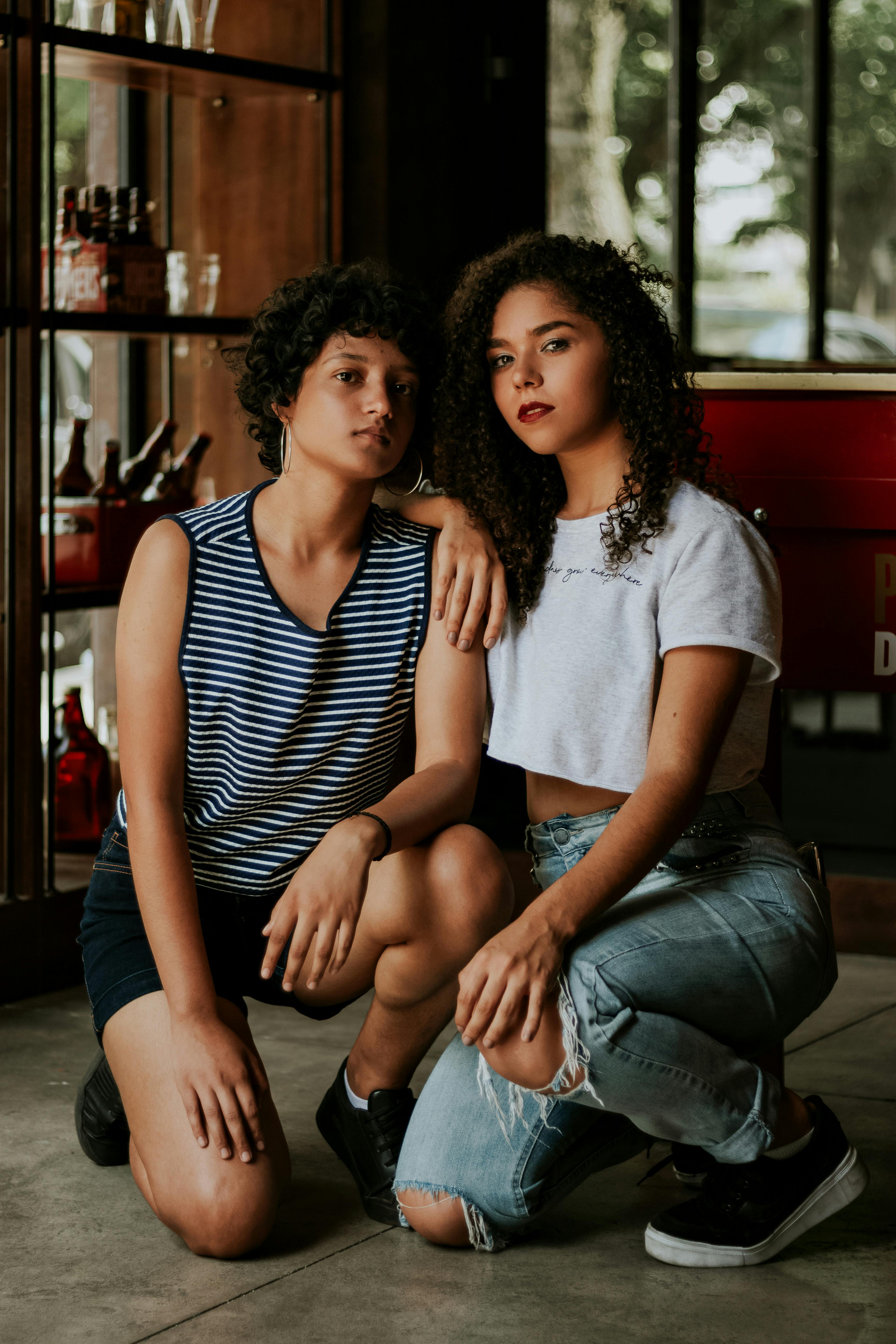 Two women posing fashionably indoors with casual attire and curly hairstyles.