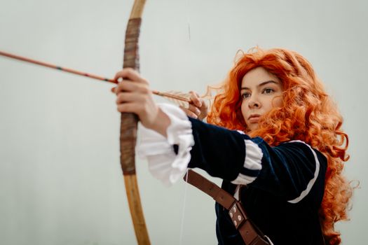 Cosplayer dressed as an archer ready to shoot an arrow, vivid orange wig and blue costume.
