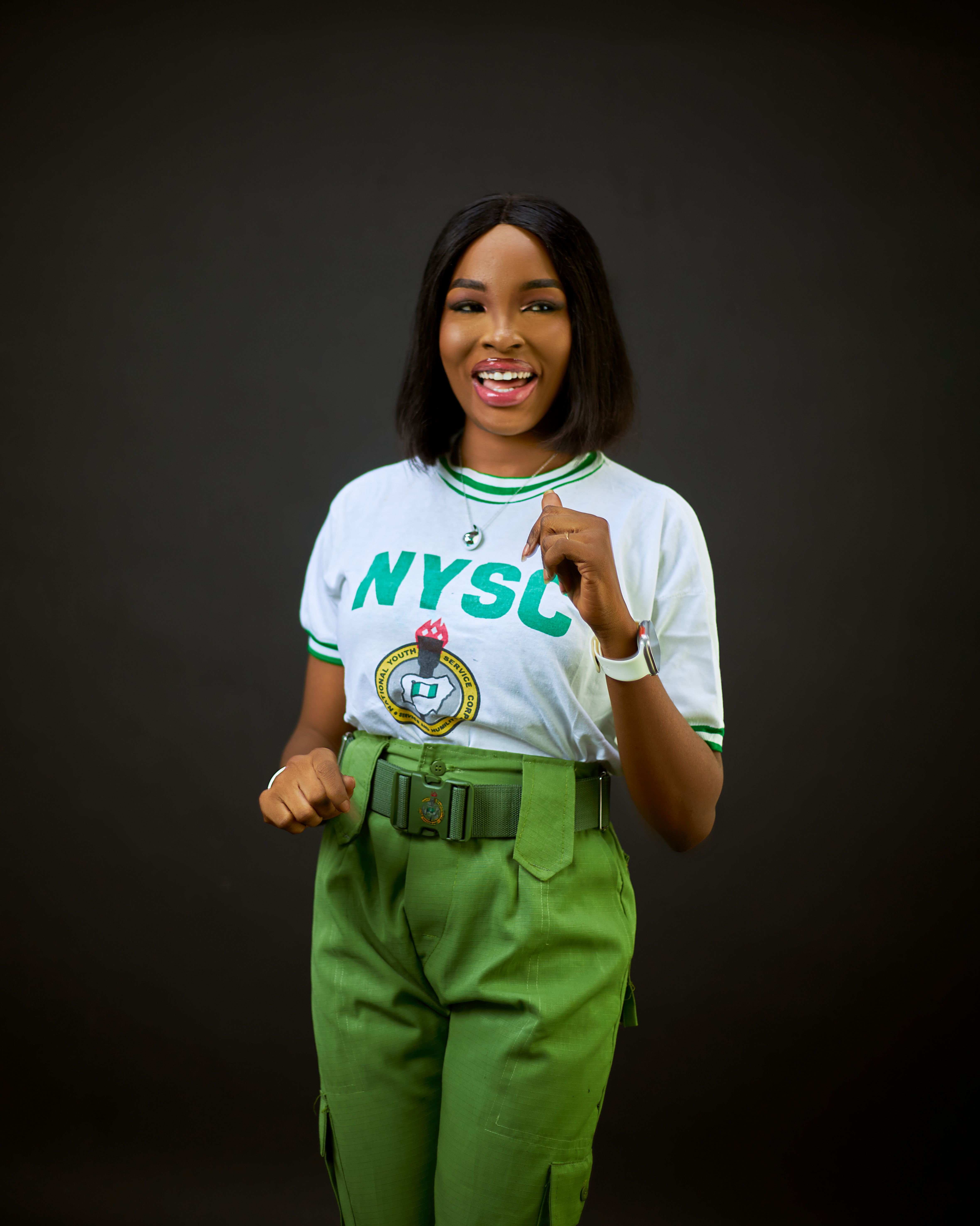 A cheerful young woman posing confidently in a NYSC uniform against a dark background.