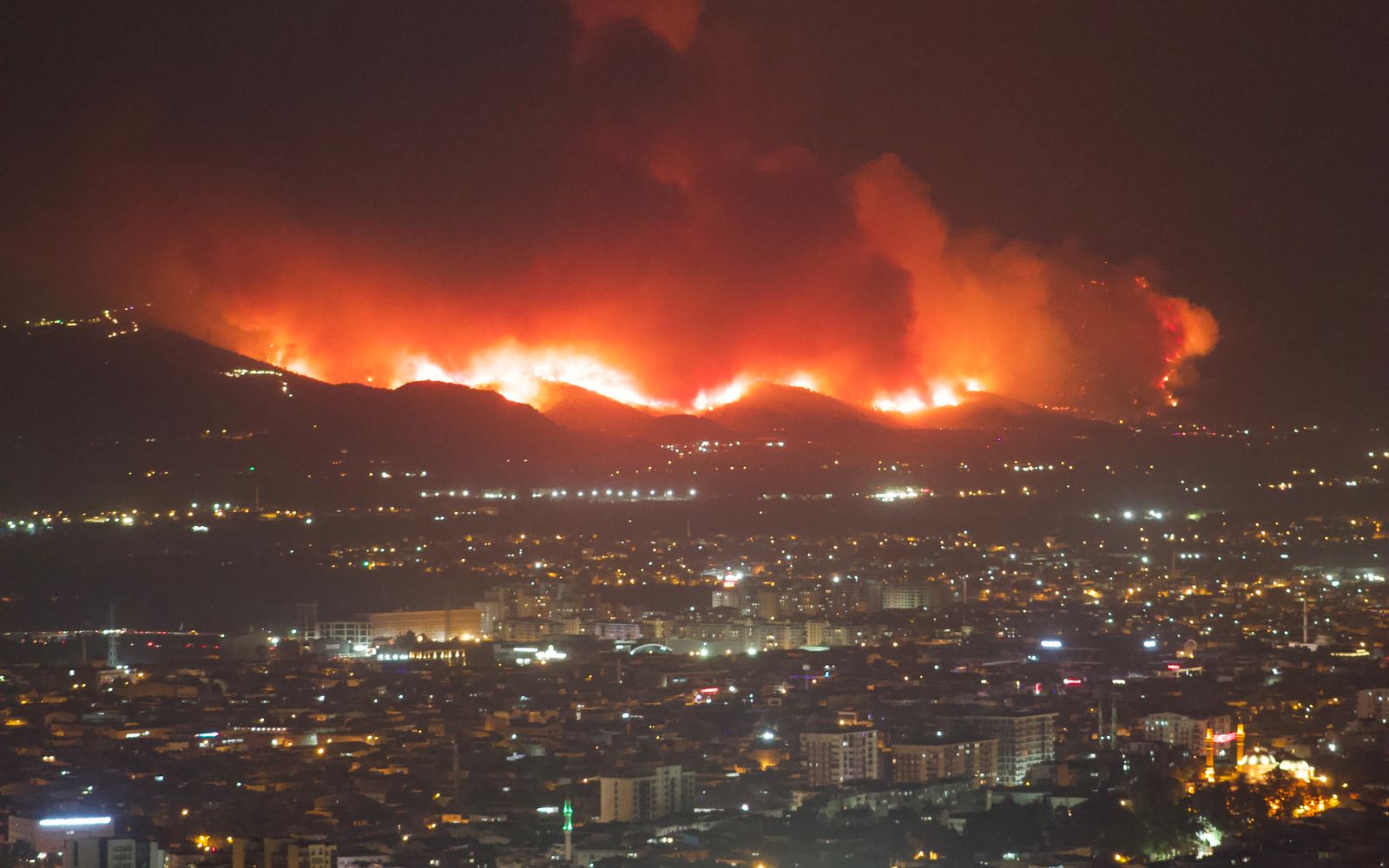 Aerial night view of wildfires burning near a city, flames lighting up the dark sky.
