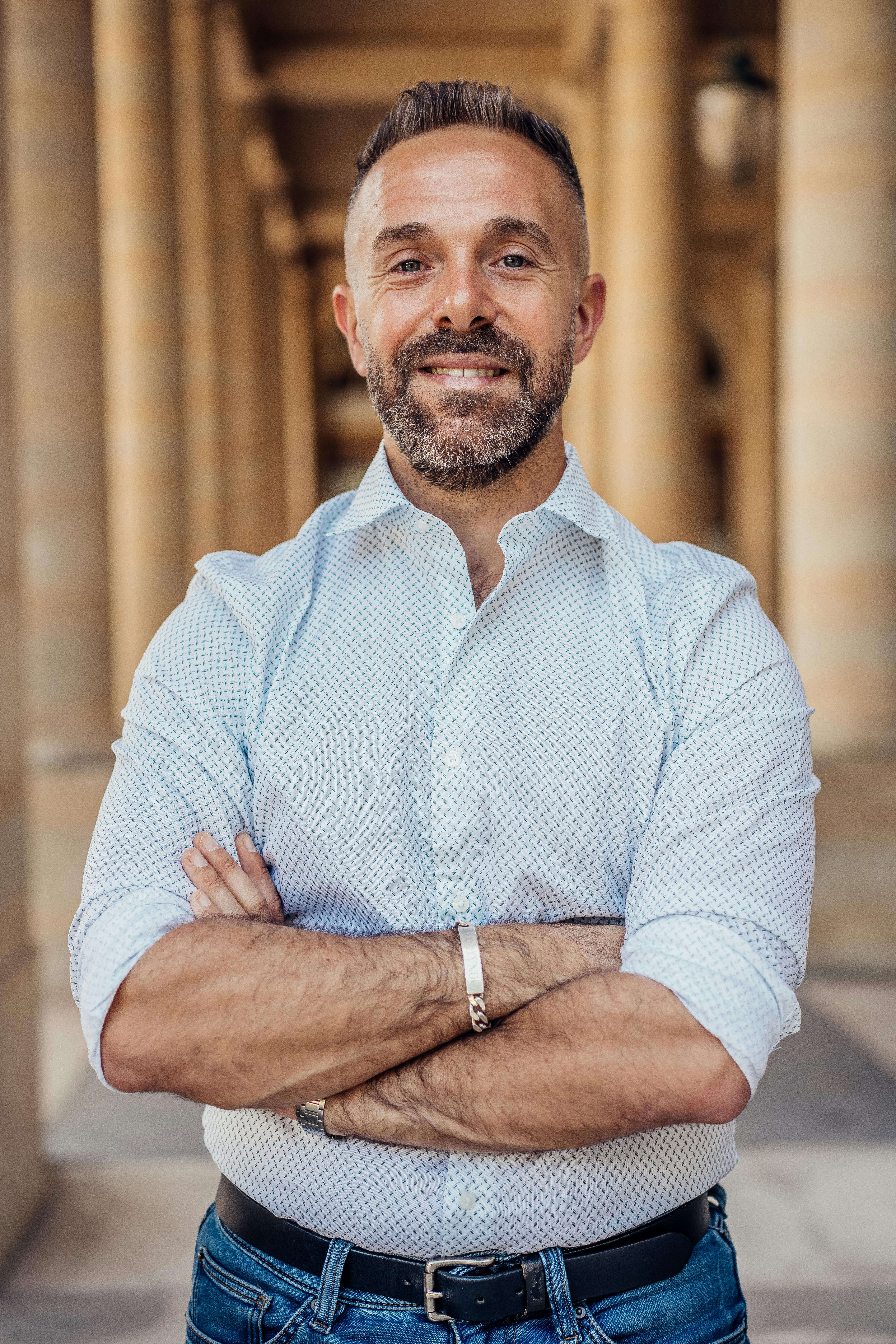 Confident man standing with crossed arms in a historic Parisian arcade, exuding professionalism and style.