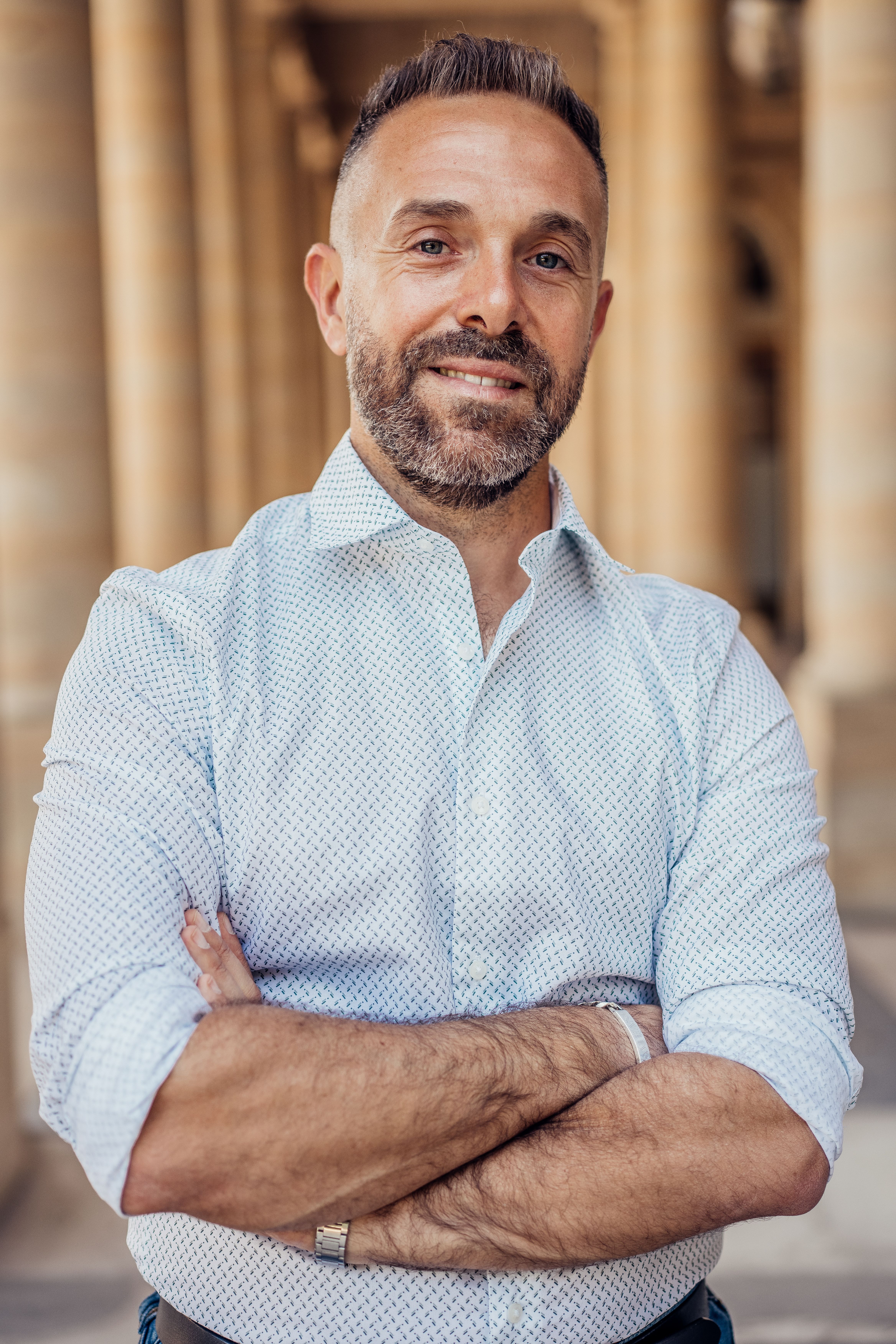 Portrait of a confident adult male in a light blue shirt, posing in a Parisian colonnade.