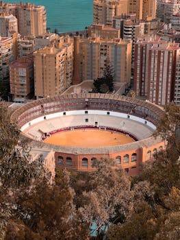 A stunning aerial view of Málaga's historic bullfighting arena surrounded by urban landscape under a warm summer sunset.